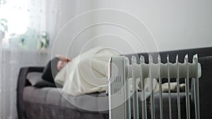 A man in a warm jacket with a hood lies on the sofa against the backdrop of a switched-on electric heater. The concept