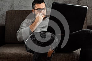 A man watches an adult video on a laptop while sitting on the couch. The concept of porn, men`s needs, pervert, lust, desire,