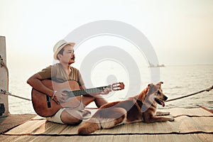 A man wear straw hat and playing guitar music song near the sea sunset with a dog pet