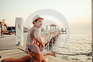 A man wear straw hat and playing guitar music song near the sea sunset with a dog pet