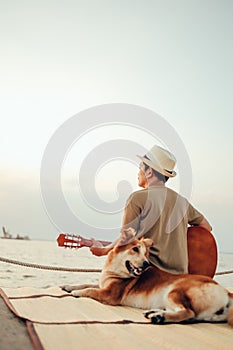 A man wear straw hat and playing guitar music song near the sea sunset with a dog pet