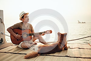 A man wear straw hat and playing guitar music song near the sea sunset with a dog pet