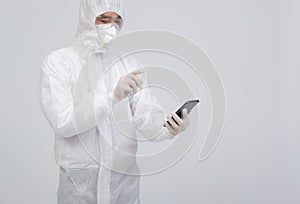 Man wearing biological protective uniform suit clothing, mask, gloves spraying sanitizer on smartphone for sanitizing virus