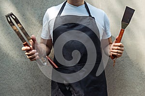 A man wearing black chef`s apron, holding barbecue tools: bbq tongs, spatula
