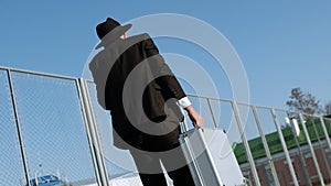 Man wearing black suit and hat with metal case in hand walks outside. Back view, behind. Businessman looks like Jew or