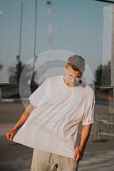 Man wearing blank t-shirt posing against glass mirror wall in the city street, front tshirt mockup on model