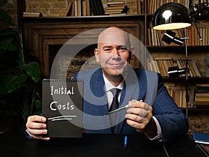 Man wearing blue business suit and showing black notepad with written text Initial Costs . Blurred background. Horizontal mockup