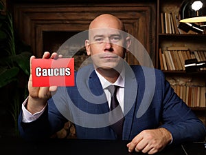 Man wearing blue business suit and showing blank red business card with written text Caucus . Blurred background. Horizontal photo