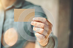 Man wearing blue jeans shirt and showing blank craft business card. Blurred background. Horizontal mockup