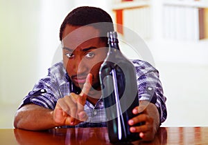 Man wearing blue white shirt sitting by bar counter holding liquor bottle and pointing waving index finger, depressed