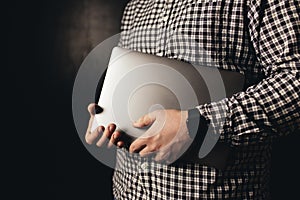 Man wearing casual clothes holds closed laptop, black blurred background