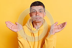 Man wearing casual style hoodie, posing isolated over yellow background, gesturing i don`t know, raising hands in bewilderment,