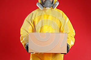 Man wearing chemical protective suit with cardboard box on background, closeup. Prevention of virus spread