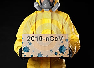 Man wearing chemical protective suit with cardboard box on black background, closeup. Coronavirus outbreak