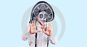 Man wearing day of the dead costume over background afraid and terrified with fear expression stop gesture with hands, shouting in