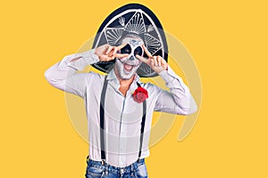 Man wearing day of the dead costume over background doing peace symbol with fingers over face, smiling cheerful showing victory