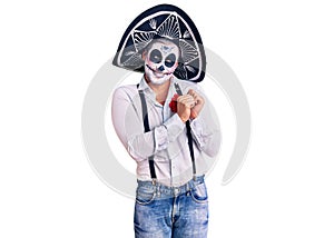 Man wearing day of the dead costume over background laughing nervous and excited with hands on chin looking to the side