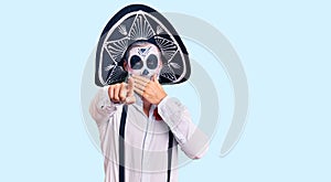 Man wearing day of the dead costume over background laughing at you, pointing finger to the camera with hand over mouth, shame