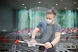 Man wearing disposable medical face mask wipes the shopping cart handle with a disinfecting cloth in supermarket. Safety during photo