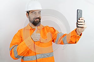 Man wearing hardhat and reflecting jacket smiling taking selfie with smartphone making thumb up sign