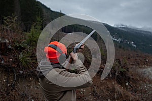 A man wearing a jacket, blaze orange hat and ear protection holds an old double barrel shotgun, aiming and shooting at orange clay