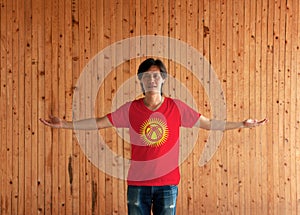 Man wearing Kyrgyzstan flag color shirt and standing with arms wide open on the wooden wall background