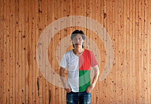 Man wearing Madagascar flag color shirt and standing with two hands in pant pockets on the wooden wall background