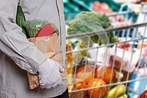 A man wearing medical gloves and a shopping bag with fresh vegetables, over blurred shopping cart with healths products. Concept