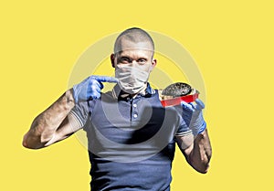 A man wearing a medical mask and gloves holds a black Burger in his hand and points at it with his index finger