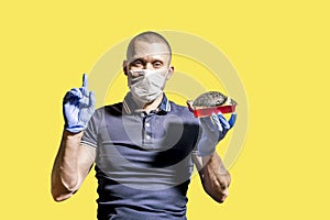 A man wearing a medical mask and gloves holds a black Burger in his hand and points with his index finger