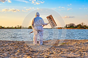 Man wearing protective suit stands with warning sign in hands with inscription closed along the sand beach