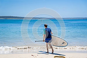 Man wearing rash guard with stand-up paddle board on a sandy beach near the ocean bay in Australia. SUP water sport activity