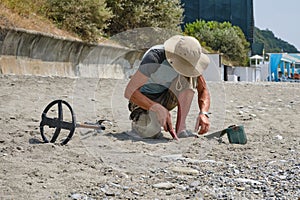 A man wearing shorts and a hat, with a metal detector, is searching for gold and coins on a sandy beach the daytime in