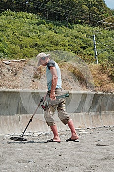 A man wearing shorts and a hat, with a metal detector, is searching for gold and coins on a sandy beach the daytime in