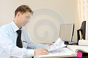 Man wearing tie sits in office of realtor and looks drawings