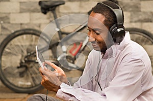 Man wearing white red business shirt sitting down, headphones on, looking at tablet screen smiling, bicycle standing