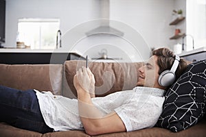 Man Wearing Wireless Headphones Lying On Sofa At Home Streaming From Mobile Phone