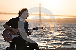 A man wearing wireless headphones sits at the quay and playing guitar at sunset - looking in the camera