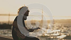 A man wearing wireless headphones sitting at the waterfront and playing electric guitar at sunset