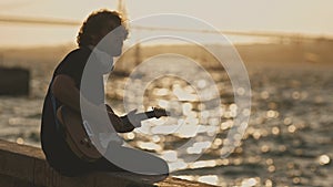 A man wearing wireless headphones sitting at the waterfront and playing guitar at sunset