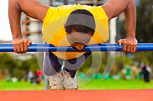 Man wearing yellow shirt and blue shorts doing static strength excercises hanging from pole, outdoors training facility