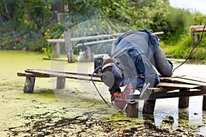 Man welder in blue uniform works, welding mask and protective red gauntlets, wooden bridge in the swamp.