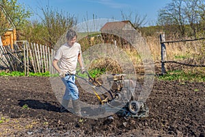 Man in wellingtons with cultivator ploughing ground in sunny day. Farmer plowing kitchen-garden in suburb. Land cultivation, soil