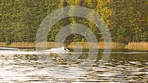 Man in wetsuit wakeboarding on serene lake, surrounded by lush green trees in peaceful forest on sunny day. Clear blue water