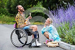 Man in wheelchair outdoors playing the guitar for blond woman sitting at curb