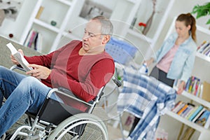 Man in wheelchair reading book carer ironing in background