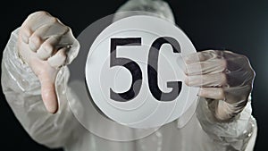 Man in white antiviral protective suit and rubber gloves holds 5G tablet and shows thumb down