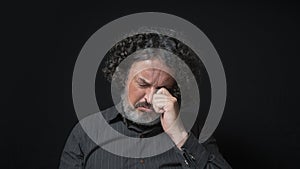 Man with white beard and black curly hair with sad expression, eyes closed and head down, wearing black shirt against black