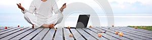 Man in white clothes meditating yoga with laptop on wooden pier