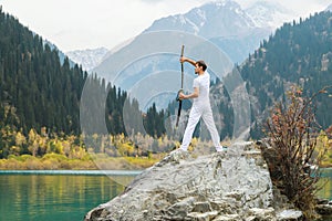 A man in white clothes with a sword stands on a large stone among the highlands